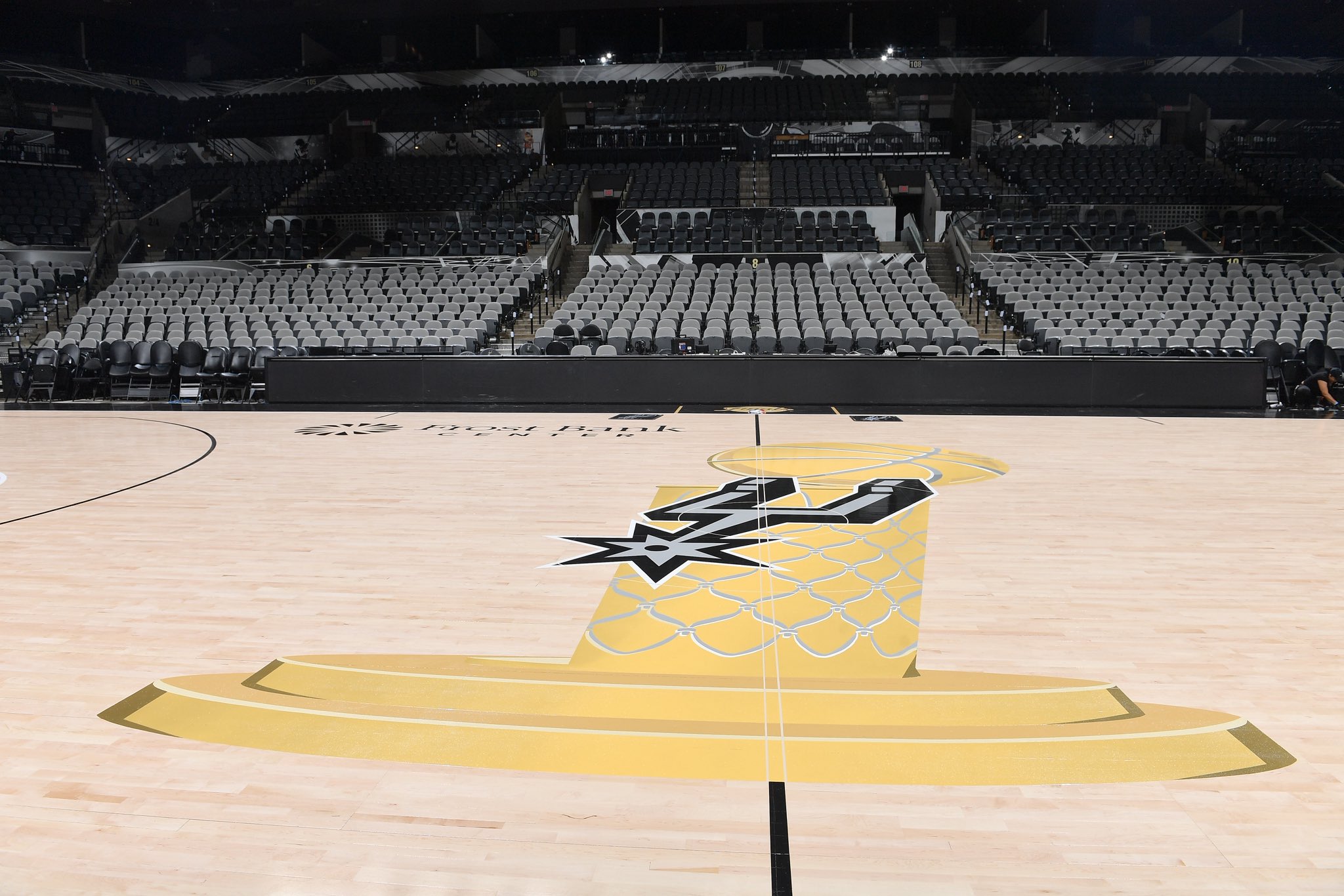 2026 NBA Finals San Antonio Spurs court at Frost Bank Center ground-level view from the baseline angle showing the Larry O'Brien Trophy painted in gold at center court with the Spurs spurs primary mark in black inside the trophy silhouette, the gold trophy base catching the arena lighting, and the natural hardwood floor extending from baseline to baseline with the empty arena bowl behind