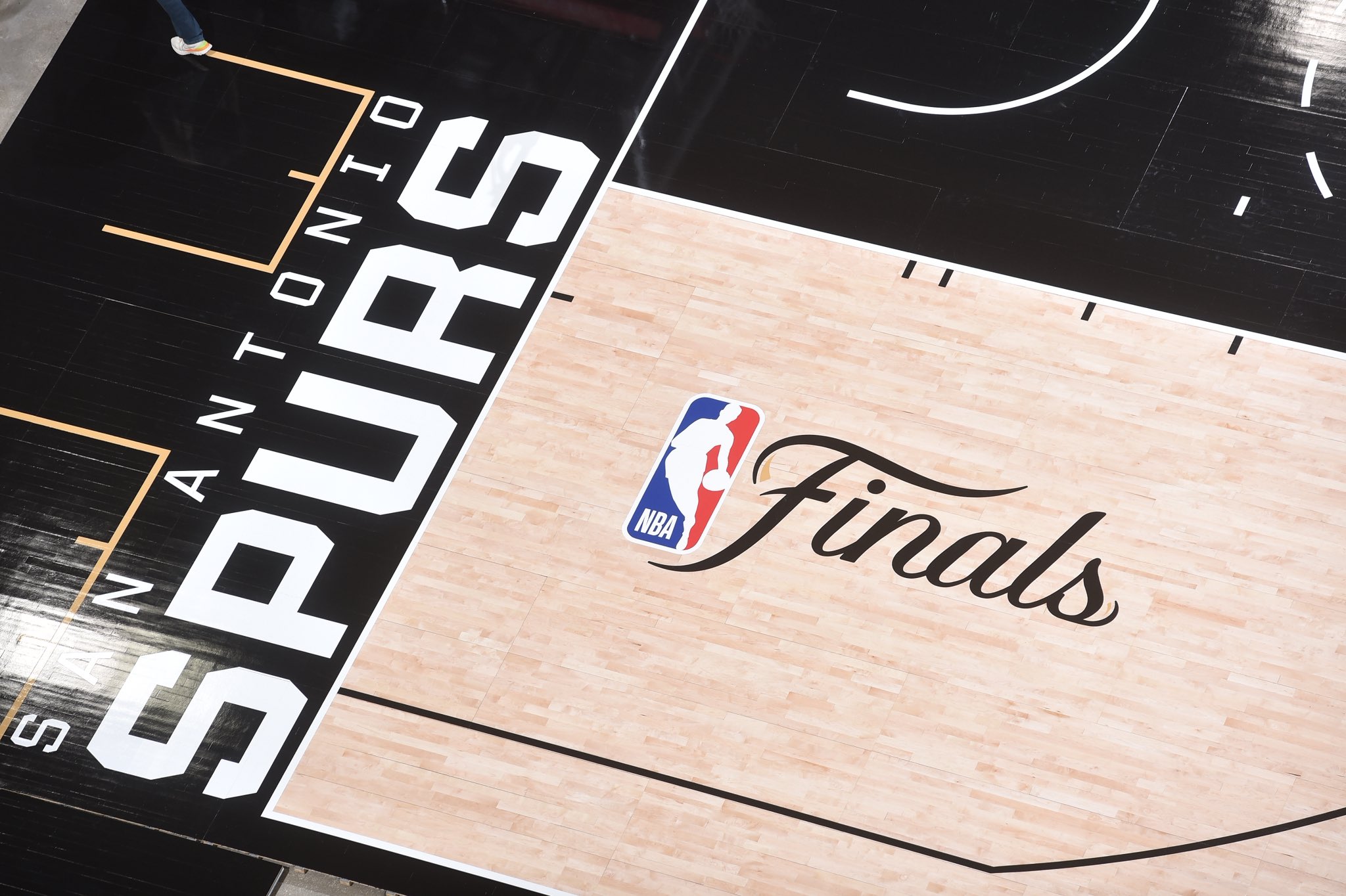2026 NBA Finals San Antonio Spurs court at Frost Bank Center close-up detail of the corner of the floor showing the SAN ANTONIO SPURS wordmark painted in white on the black border that wraps around the natural hardwood, the new NBA Finals primary logo featuring the NBA Jerry West silhouette next to the Finals cursive script, and the contrast between the black painted border and the natural wood inside the lines