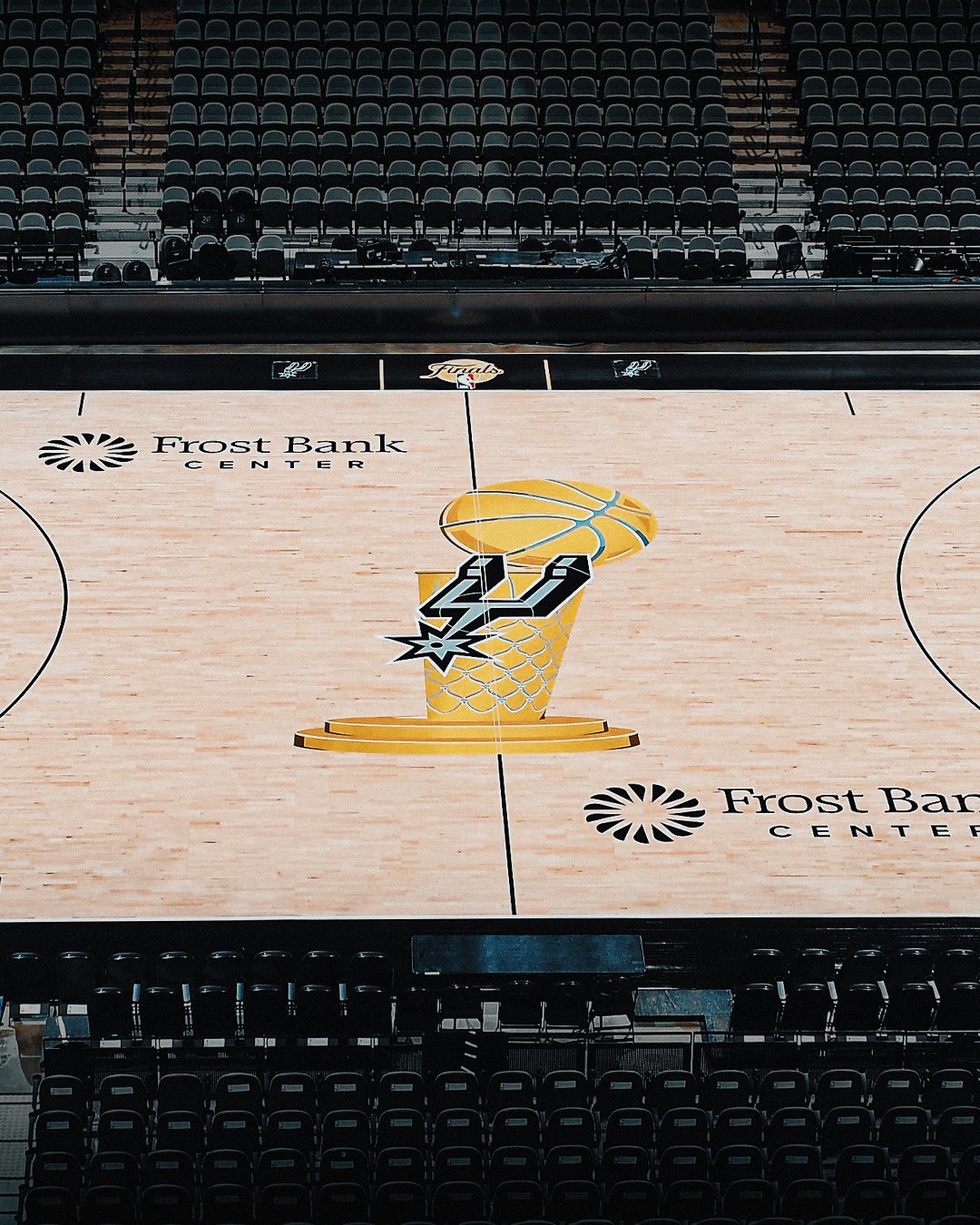 2026 NBA Finals San Antonio Spurs court at Frost Bank Center wide overhead view of the natural hardwood floor with the Larry O'Brien Trophy painted in gold at center court with the Spurs spurs mark inside the trophy silhouette, the Frost Bank Center sponsor logos above and below the trophy, and the new NBA Finals primary logo painted on the sidelines flanking center court