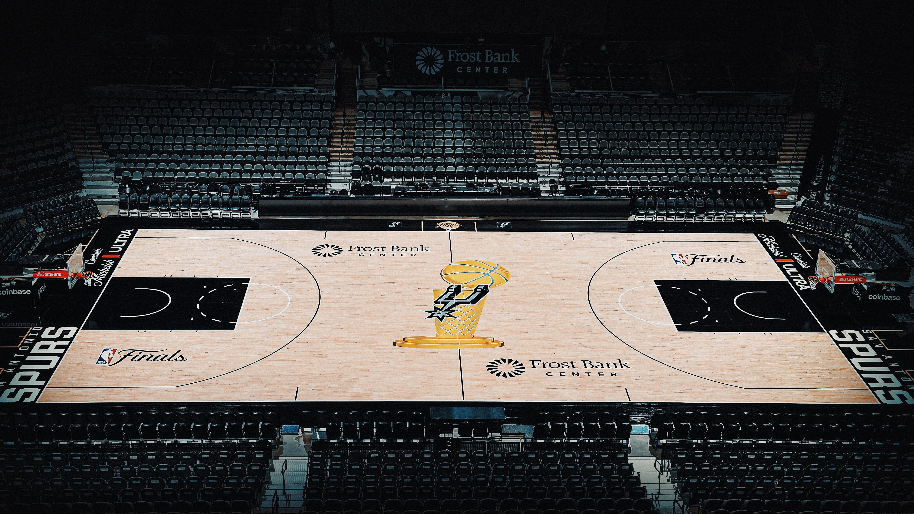 2026 NBA Finals San Antonio Spurs court at Frost Bank Center photographed in-arena from the upper bowl camera angle showing the fully painted and installed Finals floor with the Larry O'Brien Trophy in gold at center court with the Spurs primary spurs mark embedded inside the trophy silhouette, The Finals cursive script wordmark painted in both keys, the new NBA Finals primary logo painted on both sidelines directly above and below the center court trophy, Frost Bank Center sponsor logo above and below center, SAN ANTONIO SPURS wordmark in white running vertically along both baselines, Michelob Ultra and Coinbase sponsor branding on the sidelines and ad strips, and the empty arena bowl visible behind the floor