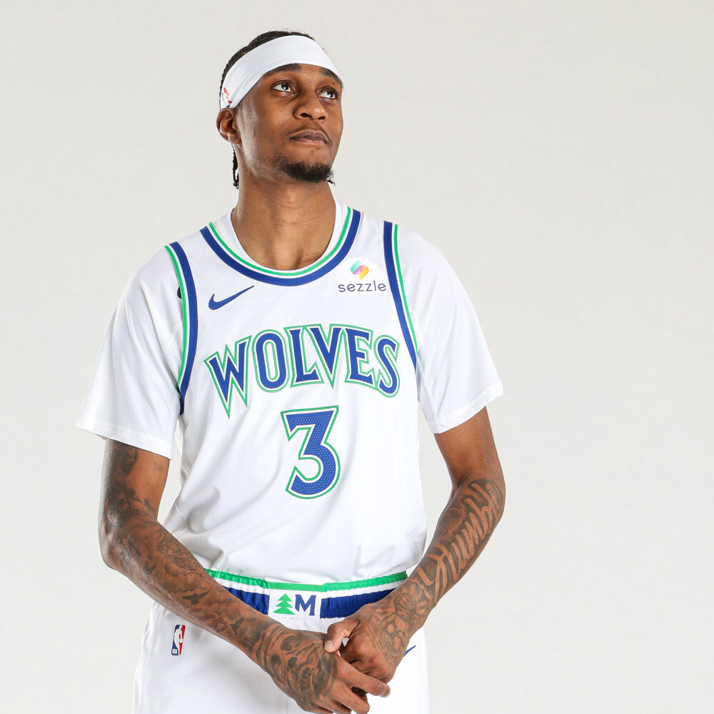 Minnesota Timberwolves new white home jersey 2026-27 front view with the throwback WOLVES wordmark in blue with green outline, brighter blue and green trim returning from the 1989 expansion identity, the Sezzle jersey patch on the chest, and the tree print pattern carrying through on the shorts waistband